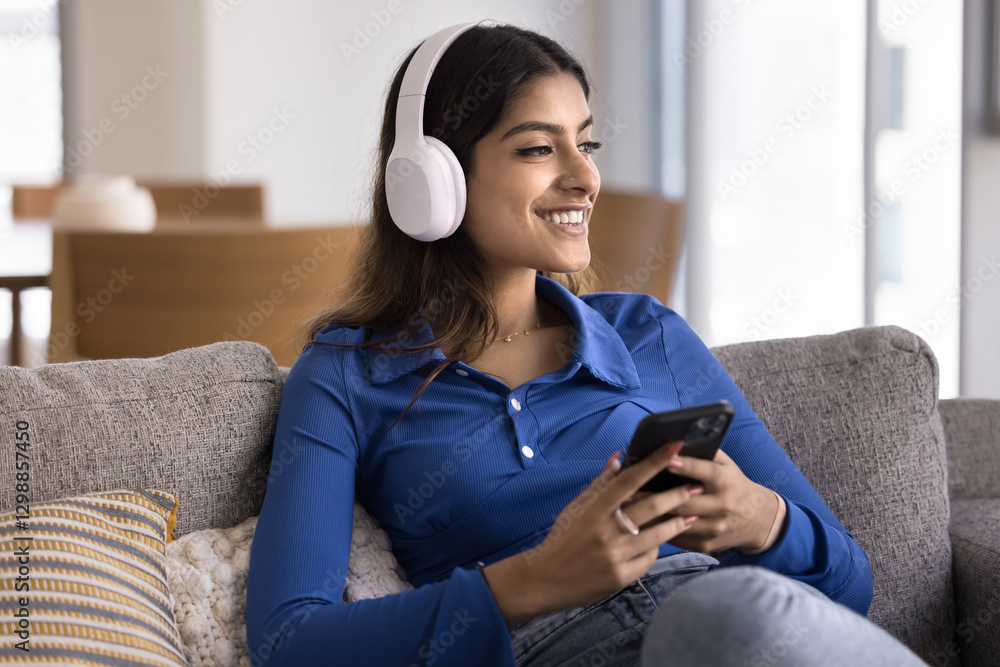 Fototapeta premium Indian woman sitting comfortably on sofa, wearing headphones, holds phone, enjoying favourite music or podcast. Relaxation through modern technology, online digital subscription services platform user