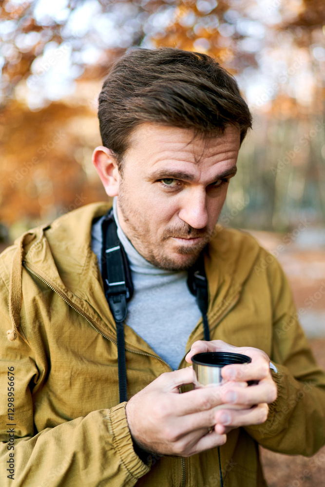 Young man with a cup of coffee in his hands looks frowningly while standing in the park Young man with a cup of coffee in his hands looks frowningly while standing in the park