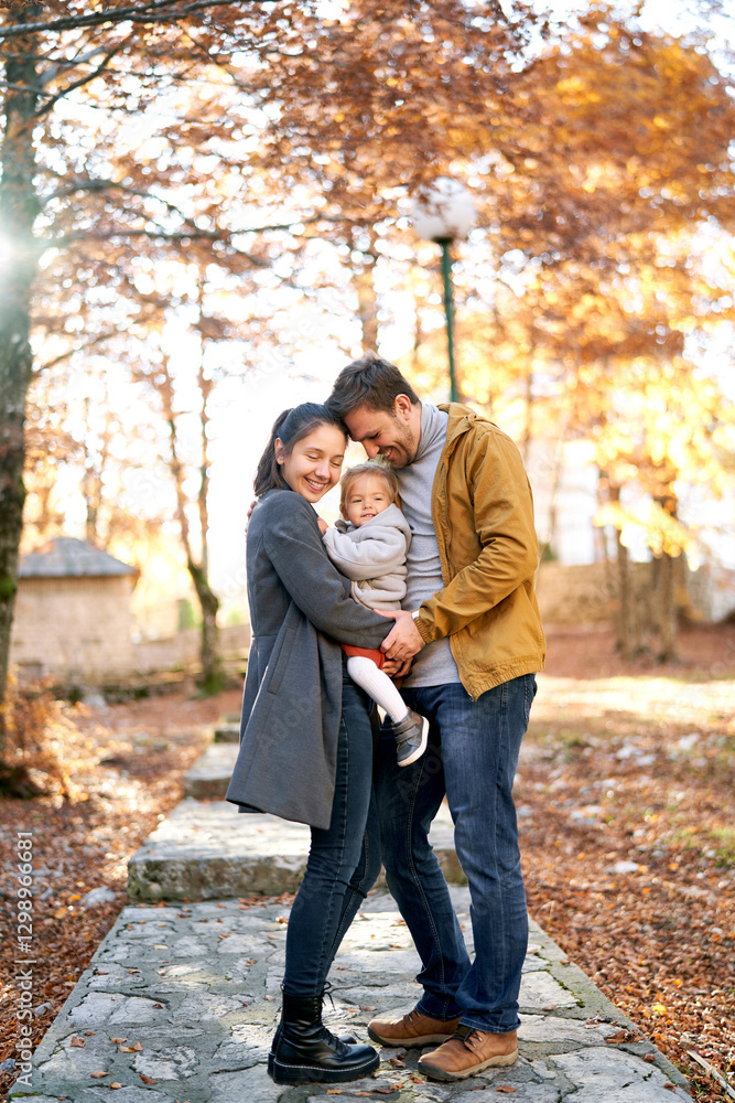 Smiling dad and mom touching foreheads while standing with a little girl in her arms in the autumn forest Smiling dad and mom touching foreheads while standing with a little girl in her arms in the autumn forest