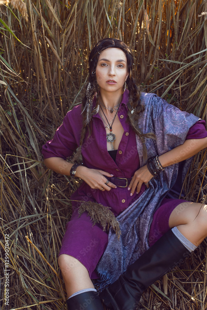 Portrait of a beautiful brunette woman in a purple dress and jewelry standing in dry bushes outside in autumn