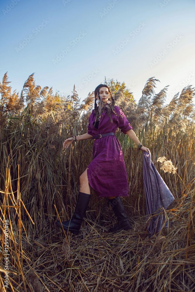 Portrait of a beautiful brunette woman in a purple dress and jewelry standing in dry bushes outside in autumn