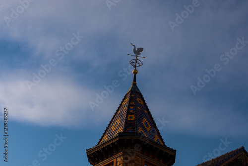 A weather vane in the shape of a swan towers on the roof spire with tiles. The sky is blue with light clouds. photo