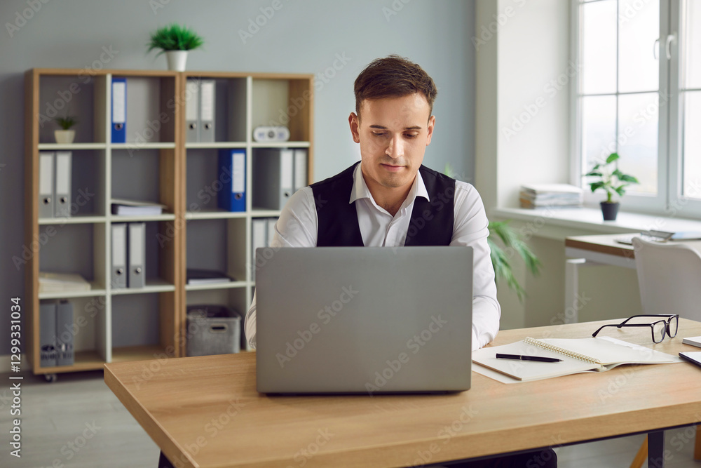 Confident young attractive business man in suit looking seriously at computer monitor screen working at the desk on his workplace and typing in the office. Male person at work with laptop. Confident young attractive business man in suit looking seriously at computer monitor screen working at the desk on his workplace and typing in the office. Male person at work with laptop.