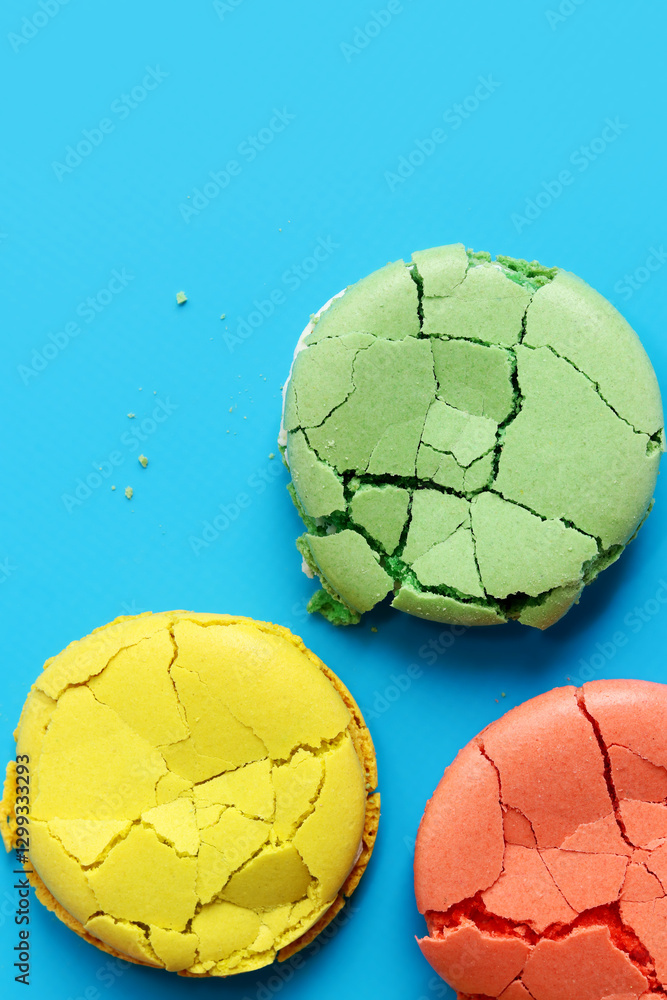 Fototapeta premium Bright multi-colored French macarons on a blue background, top view. Assortment of macarons with different flavors. Delicious pastries. Crumpled macarons, flat lay. Broken macaron on blue background