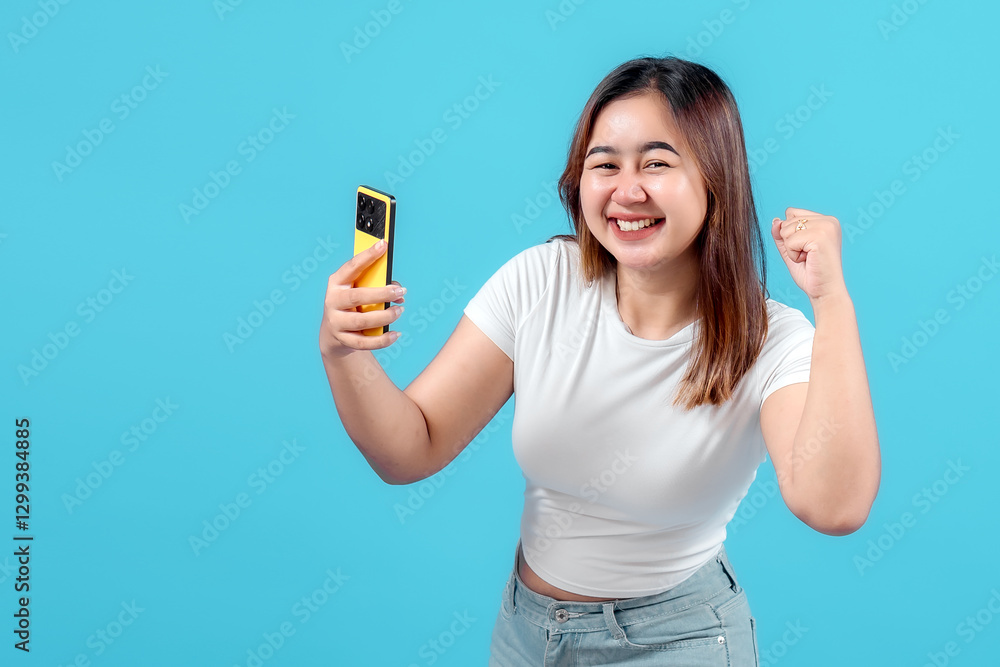 A joyful young woman celebrates while holding her smartphone, showcasing excitement and happiness against a vibrant blue background. A joyful young woman celebrates while holding her smartphone, showcasing excitement and happiness against a vibrant blue background.