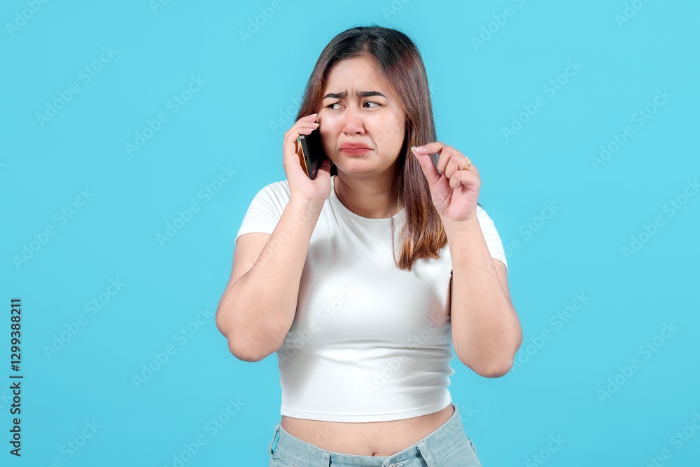 A distressed young woman cries while talking on her smartphone, expressing sadness and frustration against a bright blue background. A distressed young woman cries while talking on her smartphone, expressing sadness and frustration against a bright blue background.