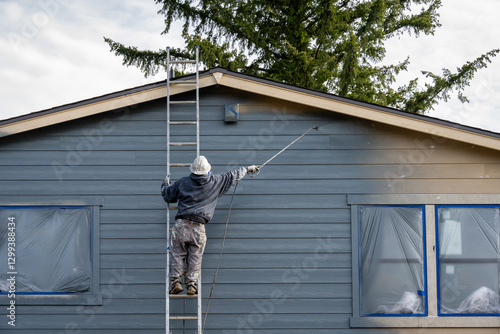 Professional painter in hard hat and air respirator on extension ladder with paint sprayer painting back exterior of house, job site for new housing development

