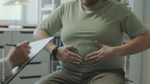 Male patient holding stomach and complaining of belly fat to female doctor taking notes on clipboard during weight loss consultation in clinic. Midsection shot
