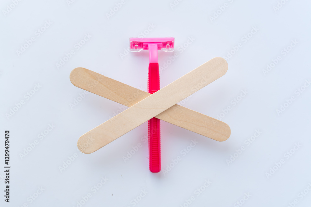 A pink razor crossed out with wooden spatulas – a symbol of rejecting shaving in favor of wax depilation. The idea of smooth skin without irritation and ingrown hairs. A pink razor crossed out with wooden spatulas – a symbol of rejecting shaving in favor of wax depilation. The idea of smooth skin without irritation and ingrown hairs.