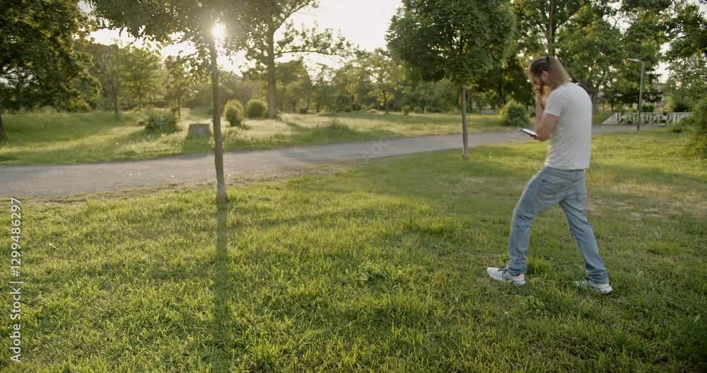A bearded man with long hair in a ponytail walks through a sunny park, talking on his phone. A bearded man with long hair in a ponytail walks through a sunny park, talking on his phone.