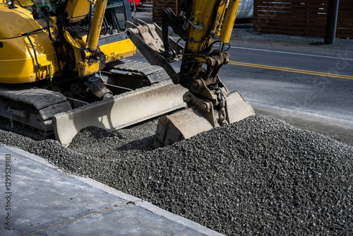 New public works sidewalk construction project between road and park, heavy equipment excavator with jawbone bucket and pile of gravel
