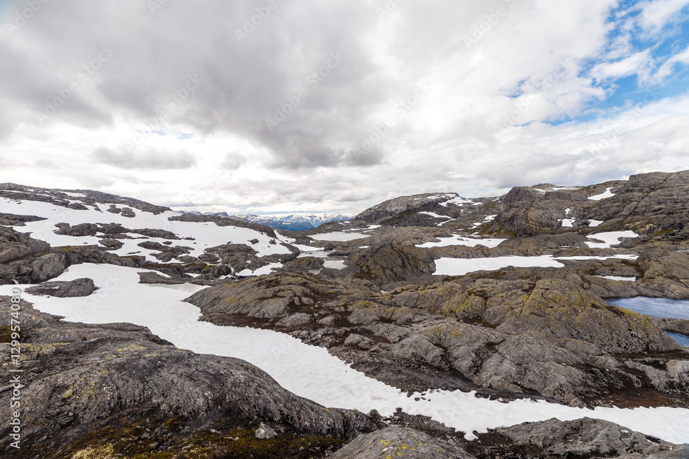 Obraz premium Stunning panoramic view of rugged terrain featuring rocky outcrops and patches of snow, under a dramatic sky filled with clouds, showcasing the beauty of nature's untouched wilderness.