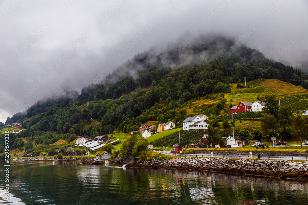 Fototapeta premium Scenic Coastal Village with Lush Green Hills and Misty Mountains Under Overcast Skies - A Serene View of Norway's Natural Beauty Along the Water's Edge