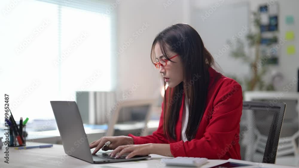 A woman wearing glasses and a red jacket is writing on a laptop. She is sitting at a desk with a chair in front of her A woman wearing glasses and a red jacket is writing on a laptop. She is sitting at a desk with a chair in front of her