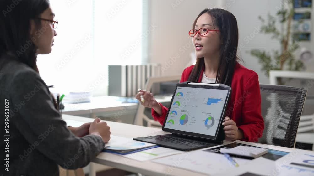 Two women are sitting at a desk with a laptop and a tablet. One of the women is holding a tablet Two women are sitting at a desk with a laptop and a tablet. One of the women is holding a tablet