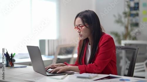 A woman is typing on a laptop computer. She is wearing glasses and a red jacket. There are several pens and a calculator on the desk
