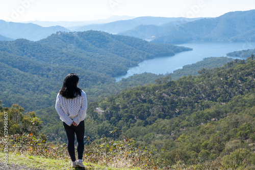 A solitary Asian female tourist stands at a scenic overlook, gazing out at a breathtaking panorama of forested mountains and Lake Eildon's skyline, Victoria, Australia. photo