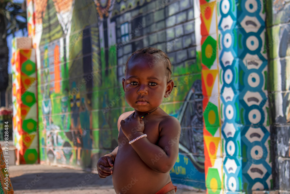 Fototapeta premium A young Namibian boy wearing traditional clothing stands next to a bright, colorful wall, his expression full of wonder.