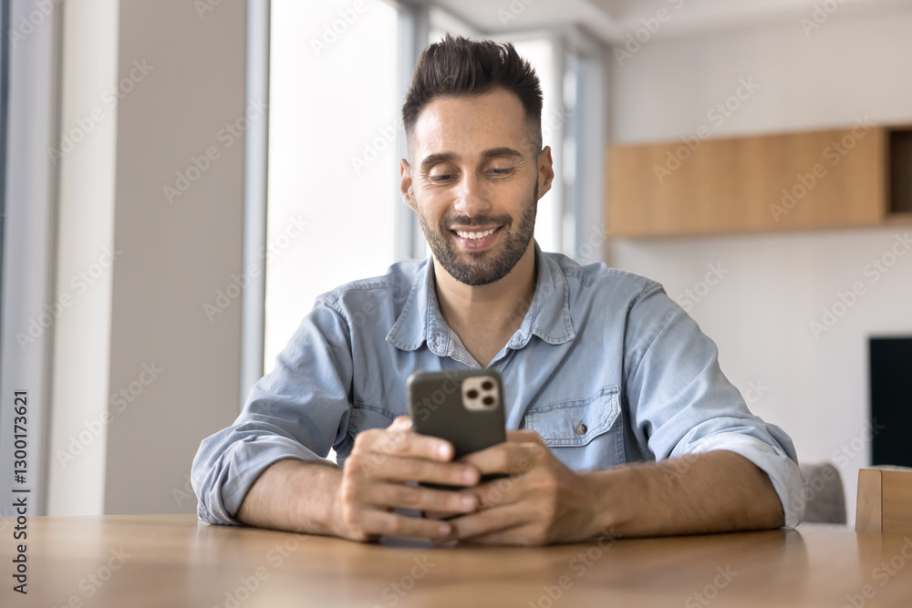 Young 30s Hispanic man using smartphone seated at table at home, smile while lead chat with girlfriend, using e-dating services mobile application, making call or buying goods though marketplace app Young 30s Hispanic man using smartphone seated at table at home, smile while lead chat with girlfriend, using e-dating services mobile application, making call or buying goods though marketplace app