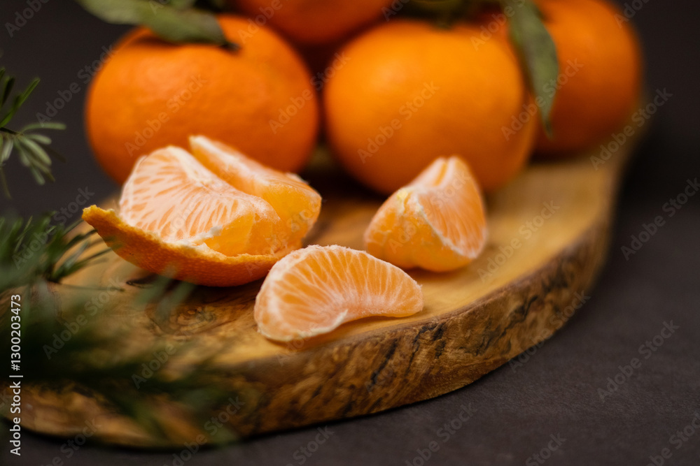 Close up of peeled mandarine slices and a pile whole mandarines on wooden plate, blurry green pine branch in front Close up of peeled mandarine slices and a pile whole mandarines on wooden plate, blurry green pine branch in front