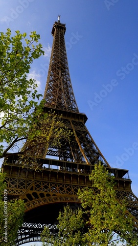 Visitors stand in awe at the base of the Eiffel Tower, admiring its intricate iron lattice against a bright sky while lush trees sway in the gentle breeze during a sunny afternoon