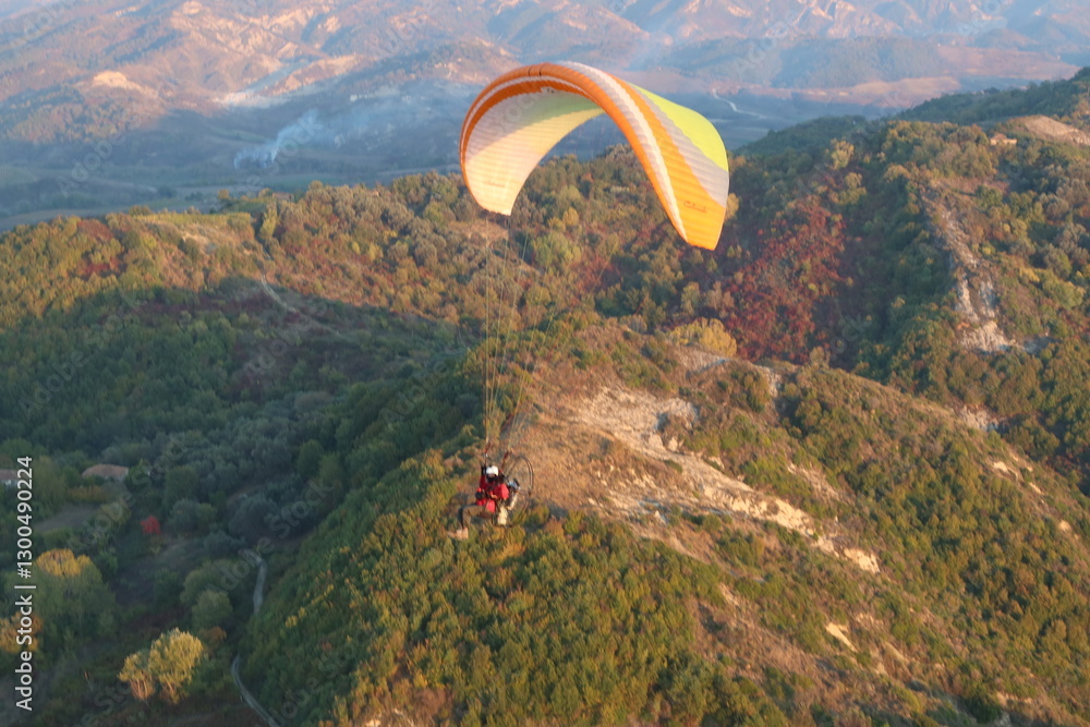 Aerial view of powered paraglider flying over mountainous landscape and forests, concept of adventure sports and aerial freedom Aerial view of powered paraglider flying over mountainous landscape and forests, concept of adventure sports and aerial freedom