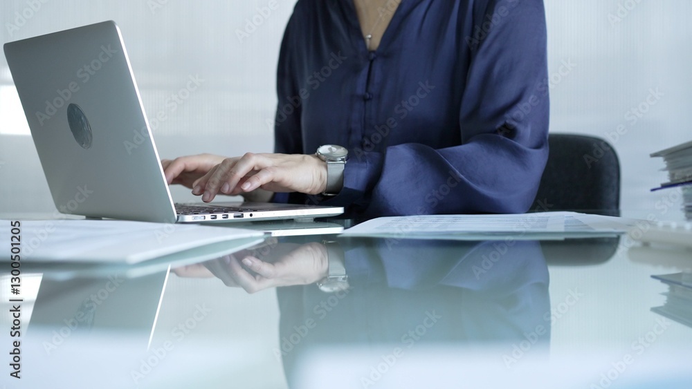 Businesswoman in blue blouse typing on laptop at glass desk while sitting in bright office setting, close-up vie. Businesspeople concept