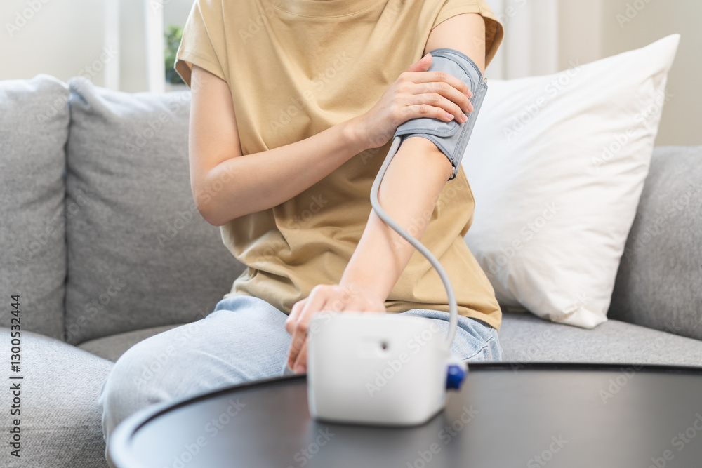 Fototapeta premium Hypotension problem asian young woman sitting on couch checking high blood pressure and heart rate with digital pressure monitor, making self check up pulse on arm, cuff at home. Healthcare medical.