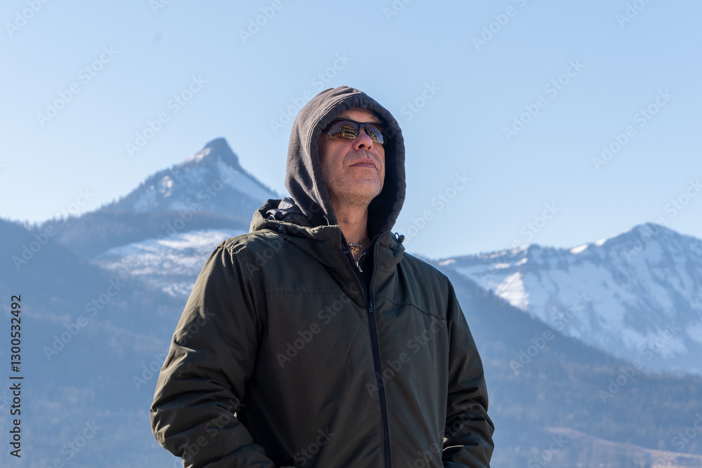 A man in a warm jacket and hoodie stands against a backdrop of snow-capped mountains. A man in a warm jacket and hoodie stands against a backdrop of snow-capped mountains.