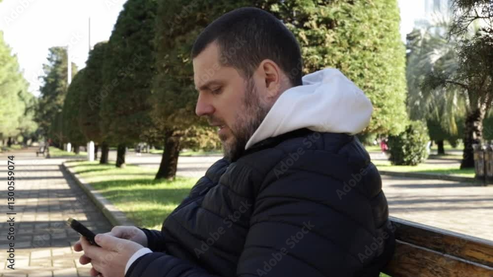 Man with phone in city park, smiling while browsing social media on his smart device, sitting on a green bench in a garden. Man with phone in city park, smiling while browsing social media on his smart device, sitting on a green bench in a garden.