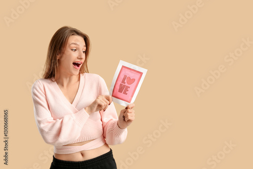 Surprised young woman holding greeting card with word LOVE and heart on beige background. Valentine's Day celebration