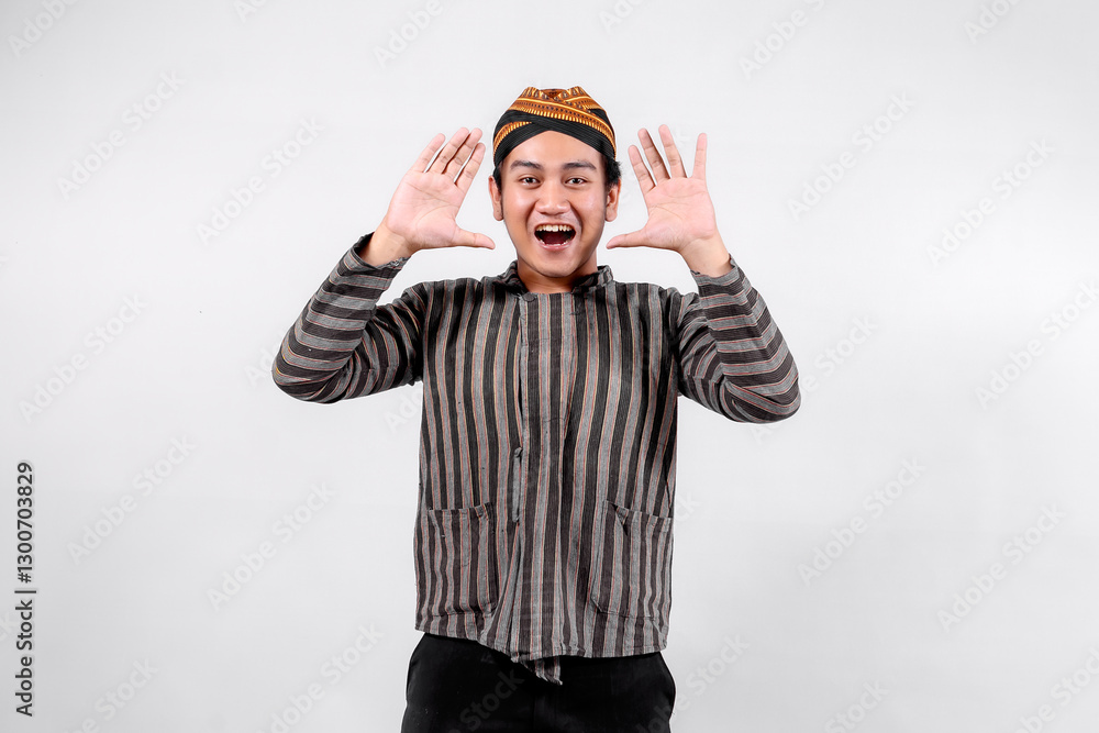 Fototapeta premium A young man wearing traditional lurik attire smiles widely while making a joyful gesture with both hands, showcasing cultural pride and enthusiasm against a neutral background.