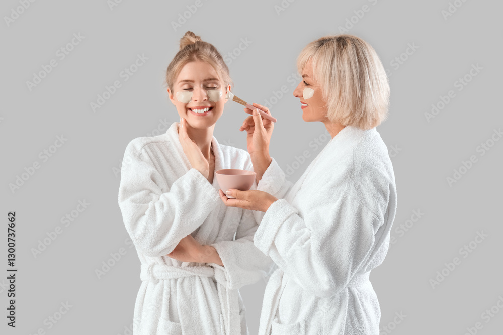 Happy young woman and her mother in bathrobes applying facial mask on grey background Happy young woman and her mother in bathrobes applying facial mask on grey background