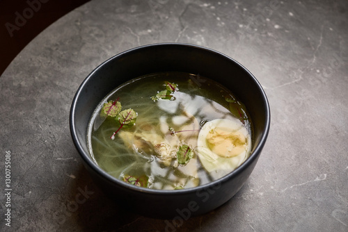 Appetizing clear soup with egg, herbs, noodles in bowl on gray marble table offering comfort and nutrition, food styling concept