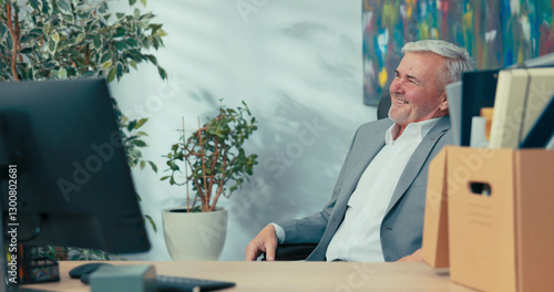 Smiling senior company executive sits in comfortable swivel chair next to computer looking around him reminiscing about years of work elegant man with gray hair spends last day before retirement
