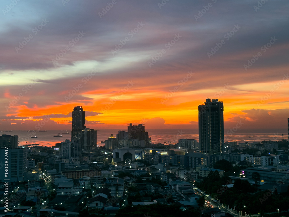 Fototapeta premium Vibrant sunset casts warm glow over coastal city skyline with tall buildings and tranquil ocean. Sriracha, Thailand.