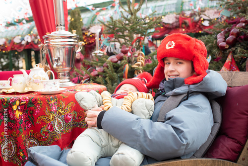 A boy in a red ushanka hat with the USSR coat of arms holds his sleeping baby brother, the baby brother has beads made of pretzels. Children sit at a table with a samovar on Maslenitsa. Pancake week i photo