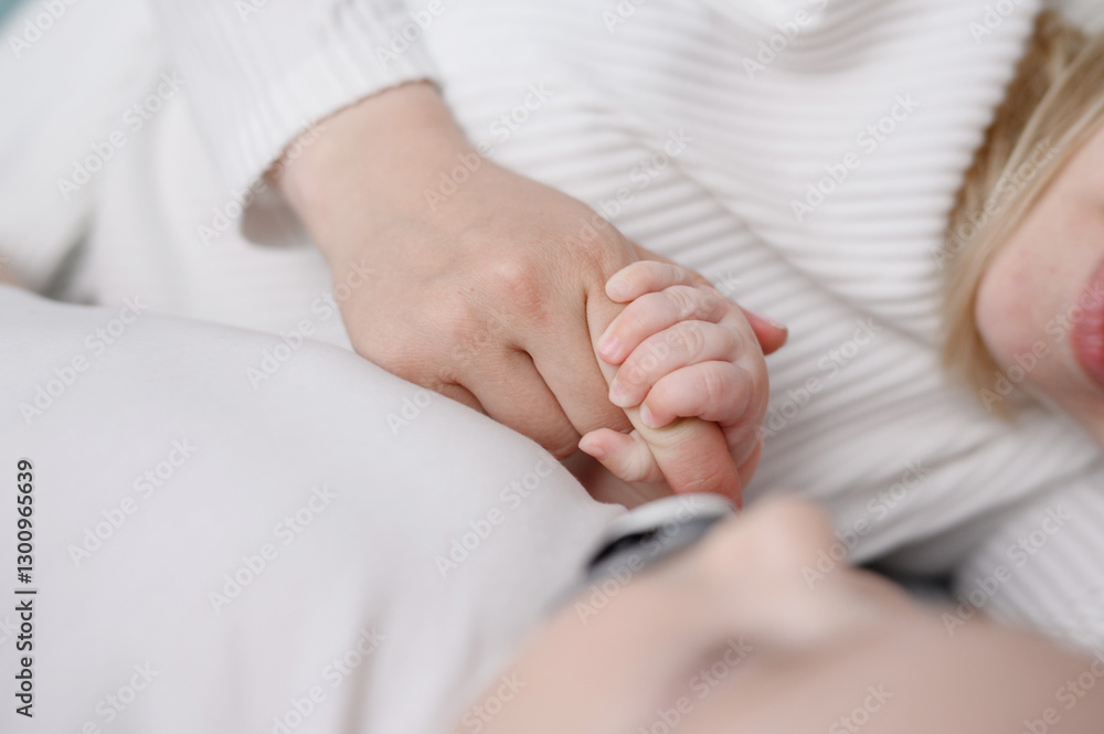 Infant lying on bed next to mom and holding mother's hand. Close up of loving mother touching little baby's hand enjoying tender moment. Motherhood and tenderness concepts Infant lying on bed next to mom and holding mother's hand. Close up of loving mother touching little baby's hand enjoying tender moment. Motherhood and tenderness concepts