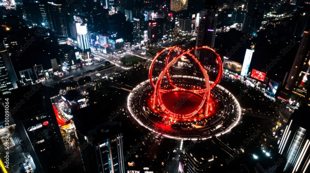 An impressive aerial night view showcasing a city centered around a stunning red circular landmark, alive with vibrant lights. An impressive aerial night view showcasing a city centered around a stunning red circular landmark, alive with vibrant lights.