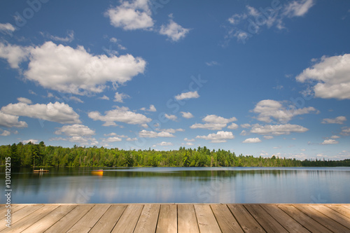 A long-exposure shot of a calm lake with a wooden dock in the foreground in Algonquin Provincial Park, Ontario, Canada. Gentle canoe movements blur on the water, with soft clouds drifting in the sky.