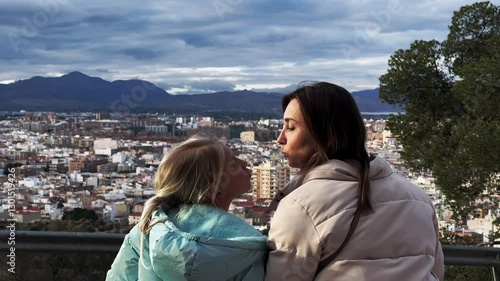 Rear view of mother and daughter kissing enjoying a beautiful city from above. Mountain range and cloudy sky in the background.