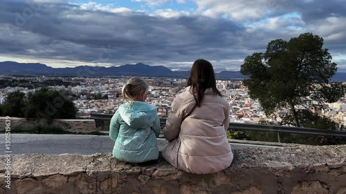 Rear view of mother and daughter tourists sitting on the parapet enjoying a beautiful city from above. Mountain range and cloudy sky in the background.