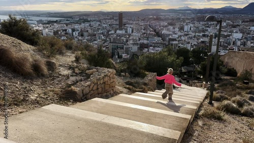 Young girl running down outdoor stairs with a cityscape in the background, captured in slow motion in sunset