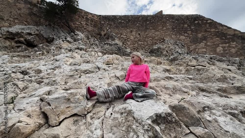 Slow motion video of a young girl wearing pink sweater and striped trousers sitting on a rocky mountain with a castle on the top.