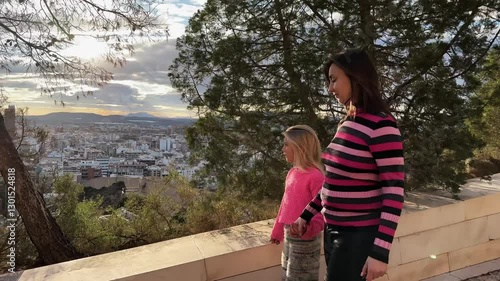 Slow motion video of a mother and daughter enjoying a walk in a park, holding hands and smiling. Sunset city in background.