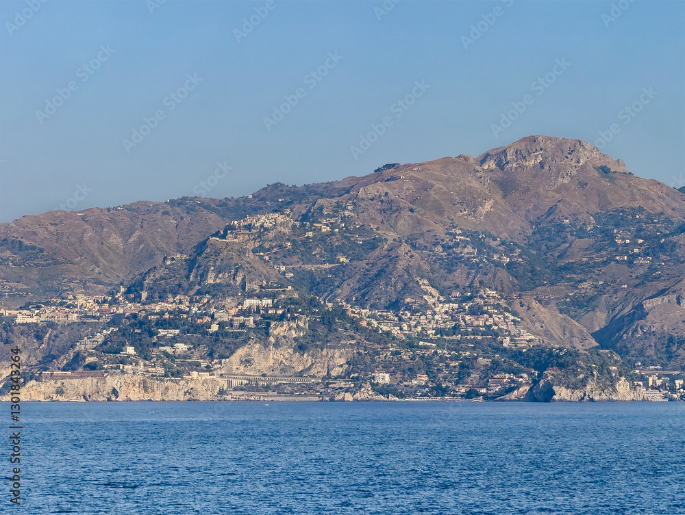 Fototapeta premium Taormina, Sicily, Italy - July 5, 2024: Taormina coastline from Isola Bella with Mendolia beach in center to the rocks of Mazzaro. Cityscape on the flank of the mountain range under blue sky