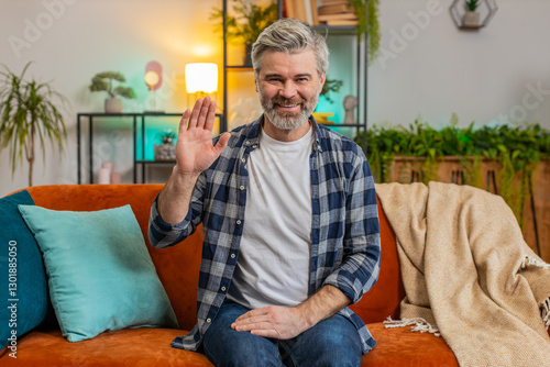 Caucasian mature man smiling friendly at camera and waving hands gesturing hello, hi, greeting, welcoming with hospitable expression at home. Senior bearded guy sitting on couch in living room at home photo