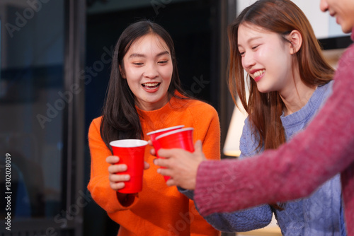 An Asian young woman with college friends sitting at a table, looking at a laptop, studying online at home, having a fun party, sharing stories, talking, and enjoying a friendly get-together
