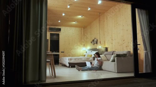 Young woman sitting on the floor, using laptop and drinking tea, working remotely from a cozy glamping pod with large windows and wooden walls at night