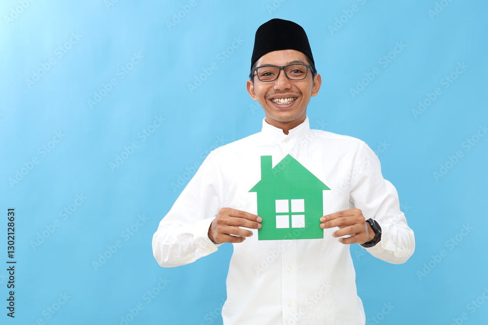 Asian muslim man holding small paper house , posing over blue color background wall in studio with copy space for promotion content or design. Asian muslim man holding small paper house , posing over blue color background wall in studio with copy space for promotion content or design.